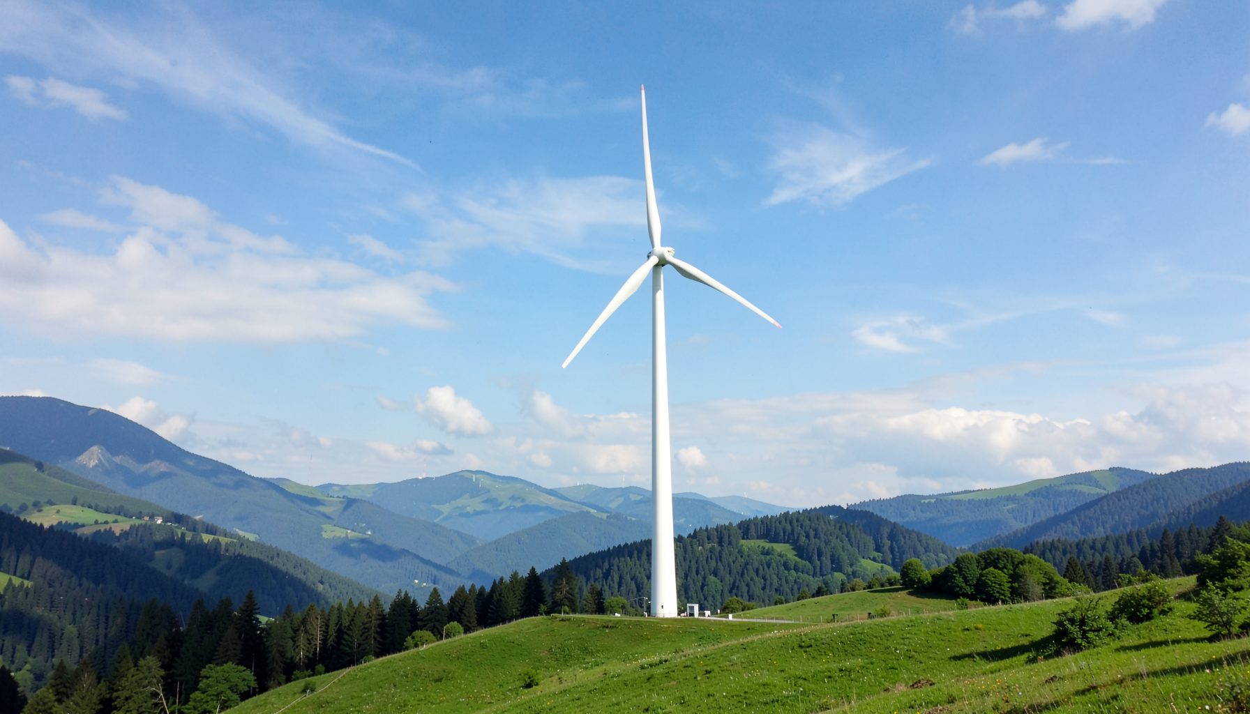 Singola turbina eolica su una collina erbosa con montagne boscose sullo sfondo e un cielo azzurro parzialmente nuvoloso.